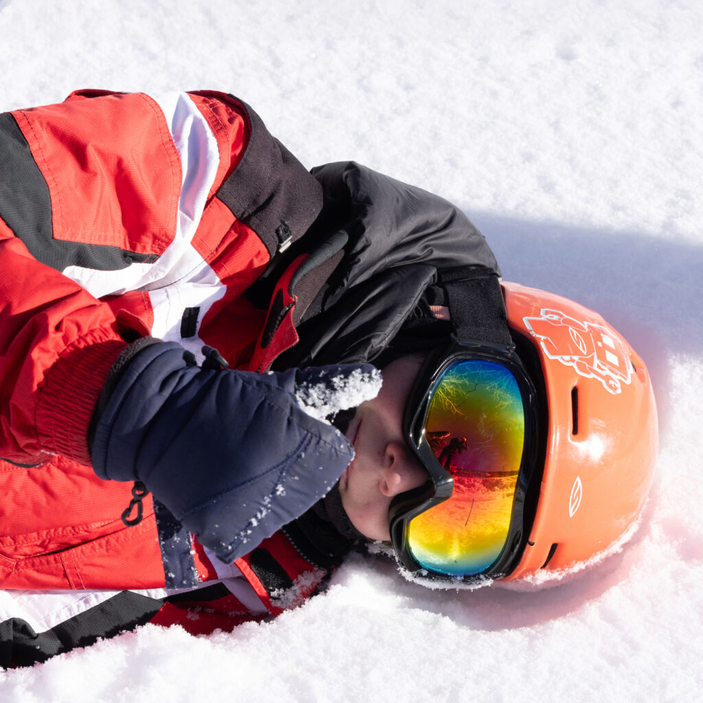 A participant wearing a helmet and goggles lies on the snow and gives a thumbs up sign during an adaptive skiing session
