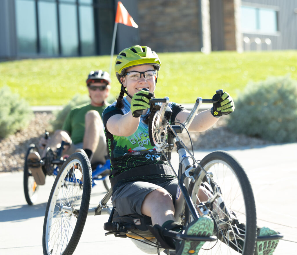 A participant uses a hand-pedaled adaptive recumbent tricycle along a paved path during an outdoor cycling program