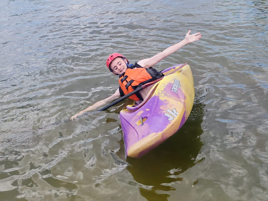 Participant wearing a helmet and life jacket practices a controlled kayak tip in calm water during an adaptive paddling activity