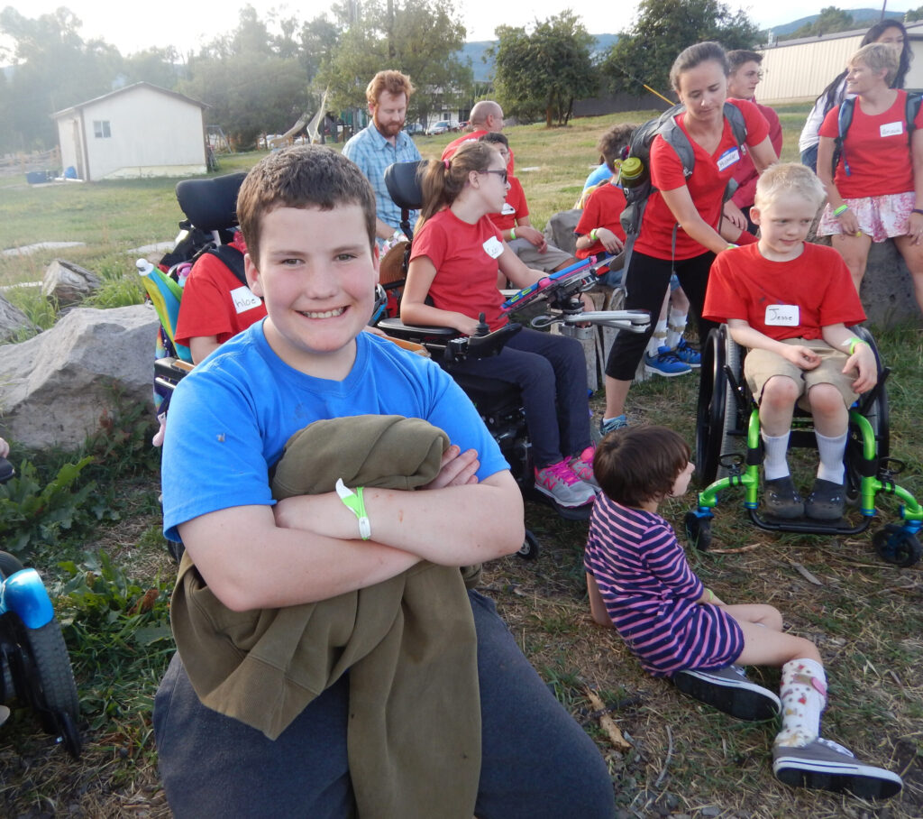A smiling participant sits in the foreground while other participants and volunteers gather together outdoors during a Summer Adventure activity