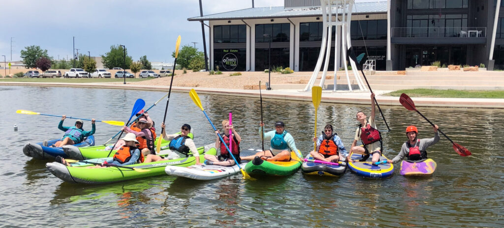 CDA participants on inflatable rafts and paddle boards line up in a row at and wave paddles in a greeting.