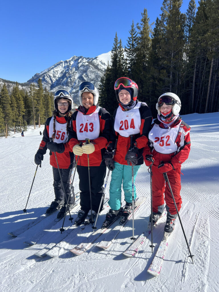 Four Team Powderhorn participants wearing race bibs stand together on a ski slope, smiling.