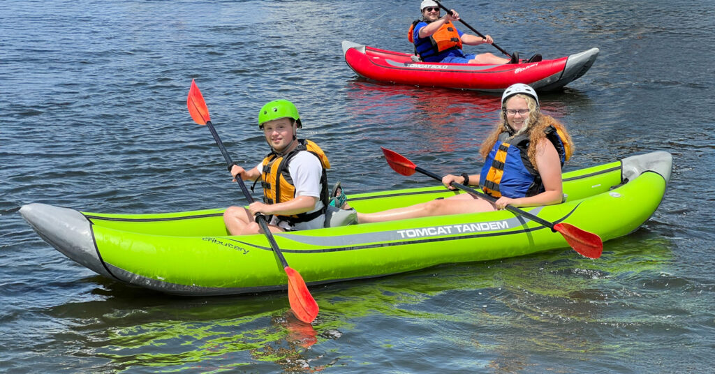 Three participants paddle inflatable kayaks on a lake during an adaptive water recreation program.