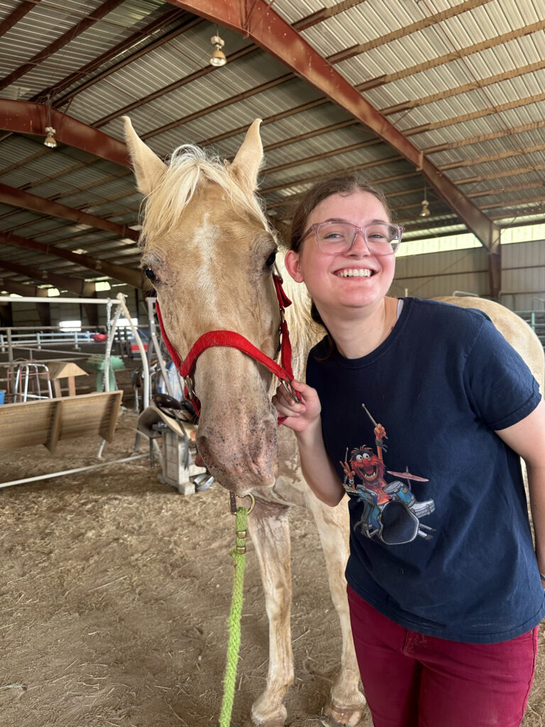 A participant smiles beside a horse inside a barn during a Summer Adventure adaptive horseback riding activity.