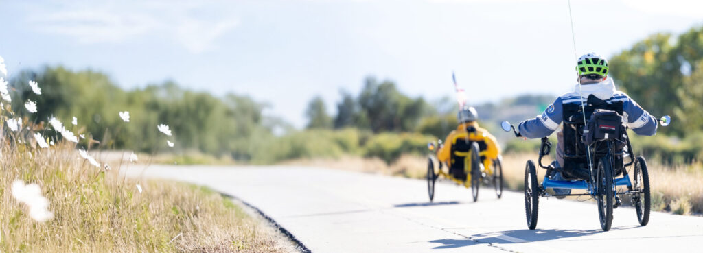 Two cycling program participants ride adaptive recumbent tricycles along the Riverfront trail.