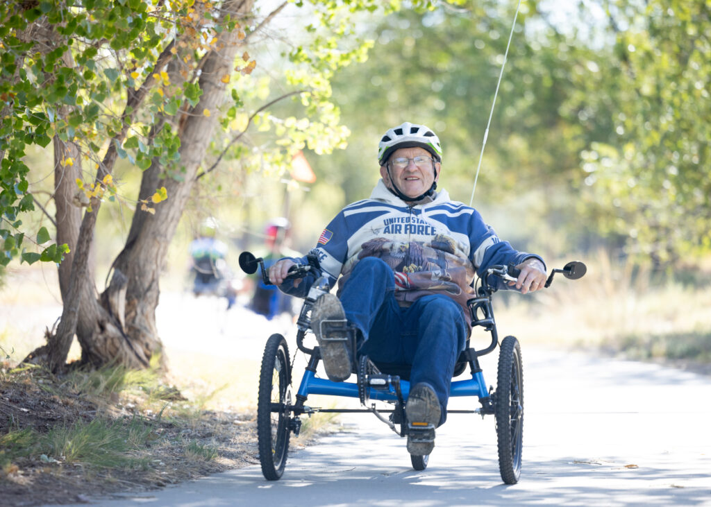 A veteran participant rides an adaptive recumbent tricycle along a paved trail during an outdoor cycling program.