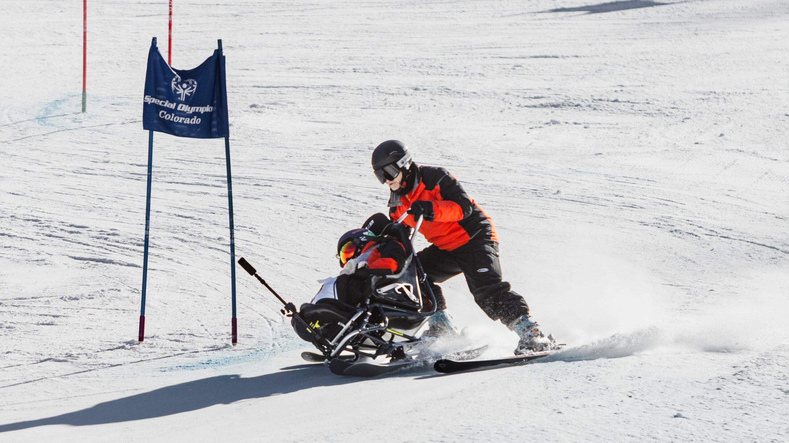 Guide skier steers a bi-ski athlete through a Special Olympics Colorado slalom gate during an alpine race.